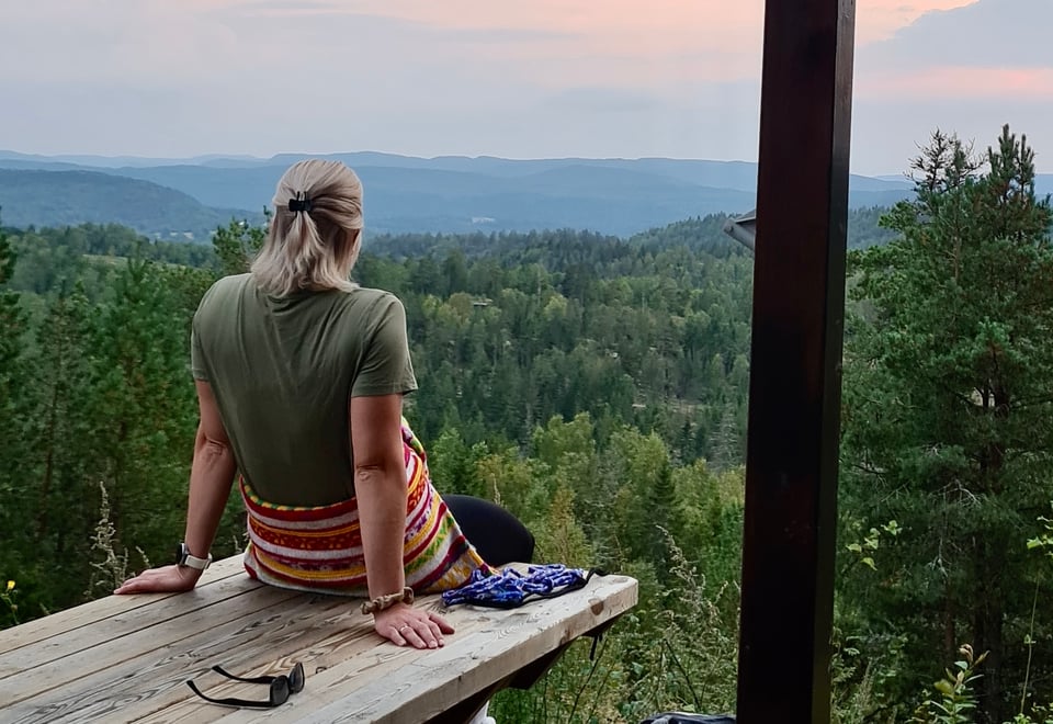 Jente sitter på en benk utenfor Tyristua i Botnemarka og ser på solnedgangen med utsikt til skog og åser.