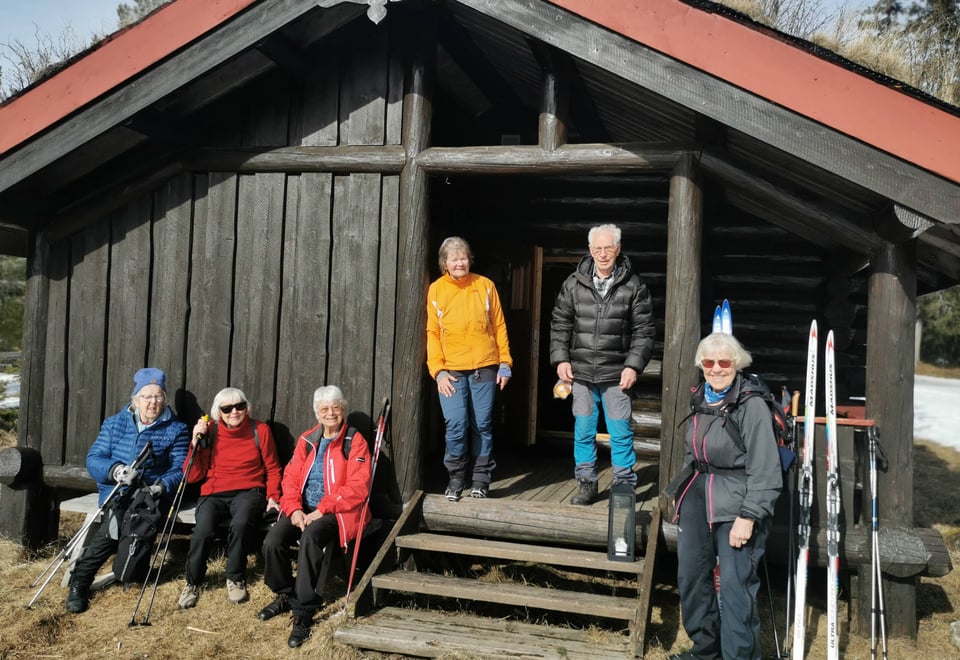 Seks eldre mennesker utenfor brun hytte.