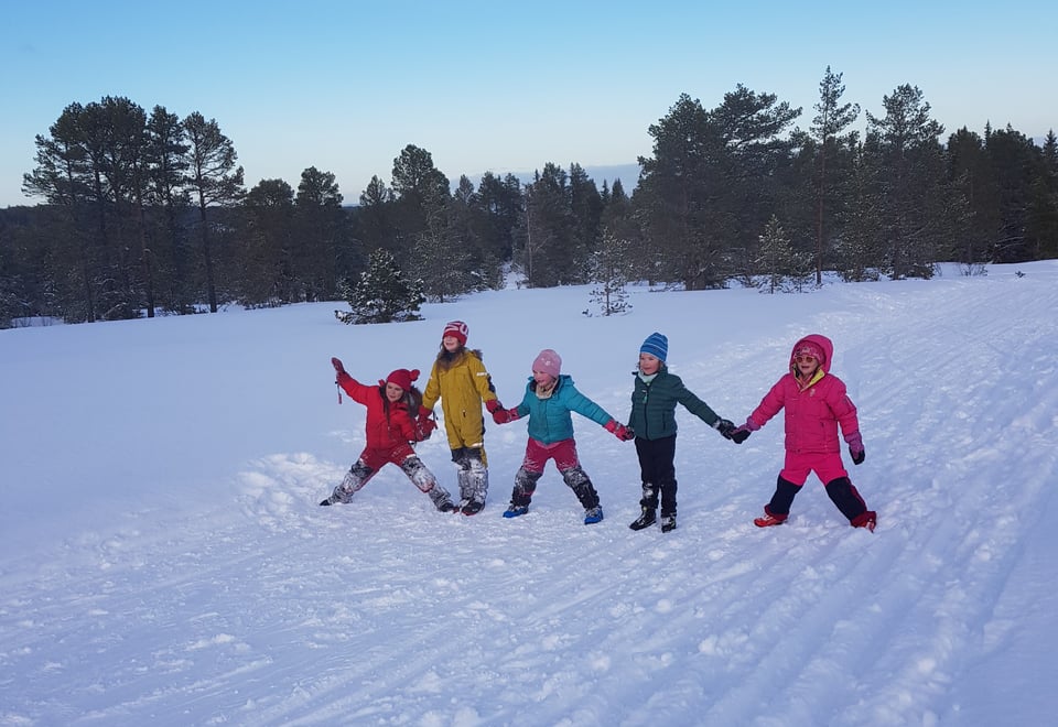 Fem barn i fargerike vinterklær som står midt i et stort felt dekket i snø, og holder hverandre i hendene i lang rekke
