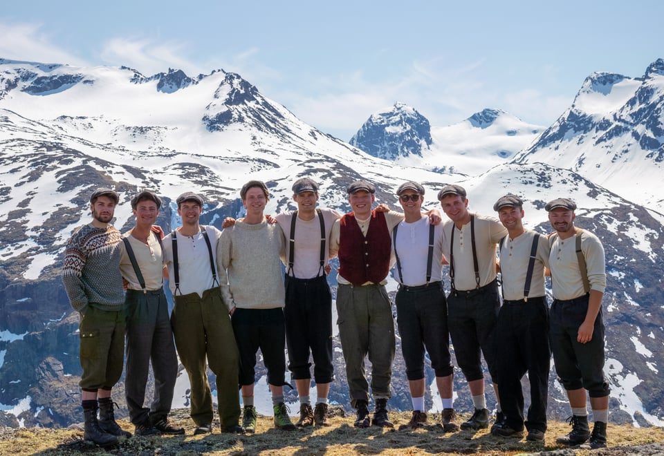 En rekke menn som står holder rundt hverandre og smiler til kamera. Bak ser vi snødekte fjell