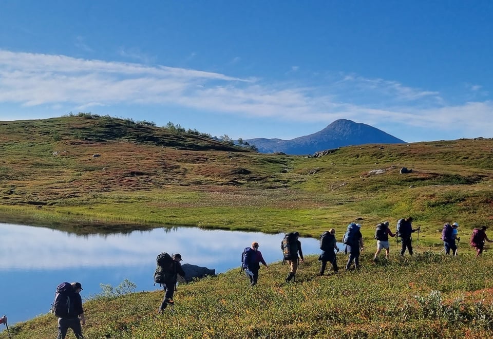 Folk med tursekk ute å går i fjellet en vakker høst dag. Blikk stille vann i bakgrunn og blå himmel.