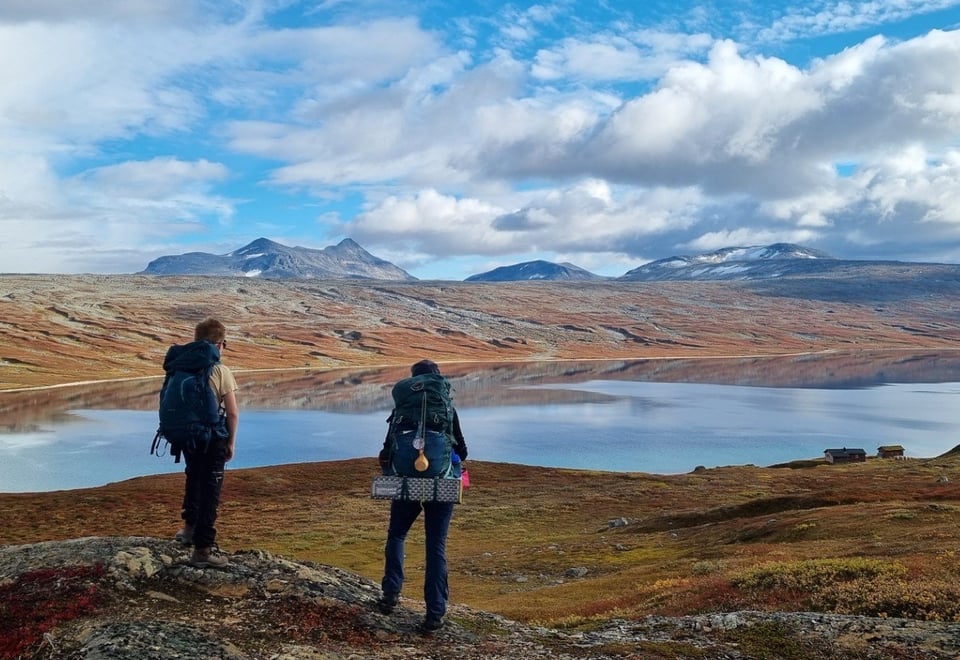 To turgåere ser ut over et fjellvatn og noen nede ved vatnet. Det er høye fjell i bakgrunnen og lette skyer