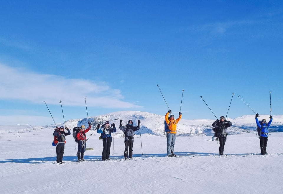Syv glade mennesker på ski i fjellet med skistavene i været. Sola skinner og det er masse snø.