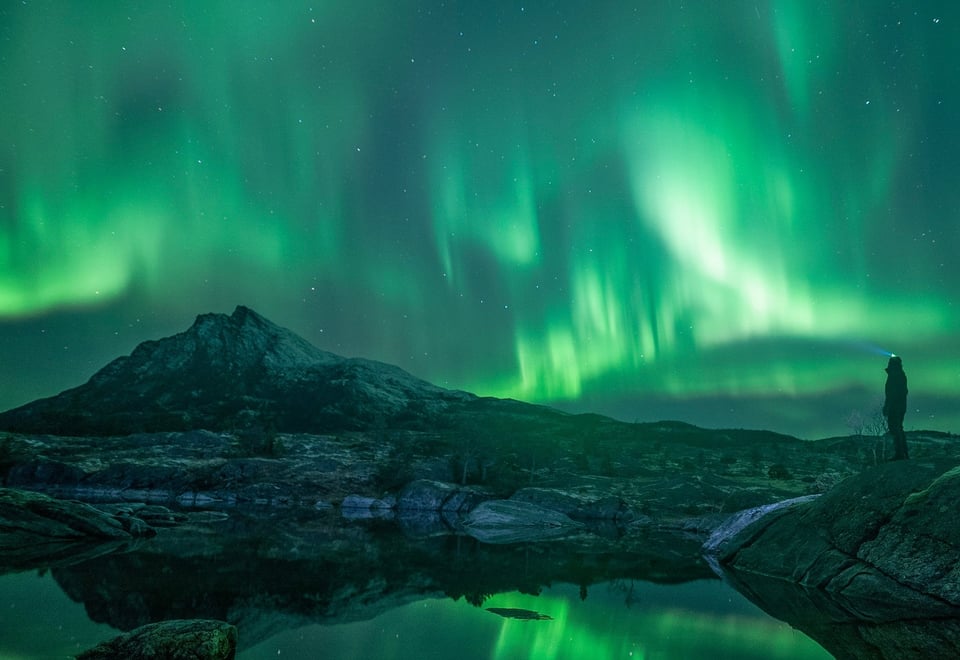 Siluetten av en person med hodelukt som står å se utover nordlyset som dekker himmelen.