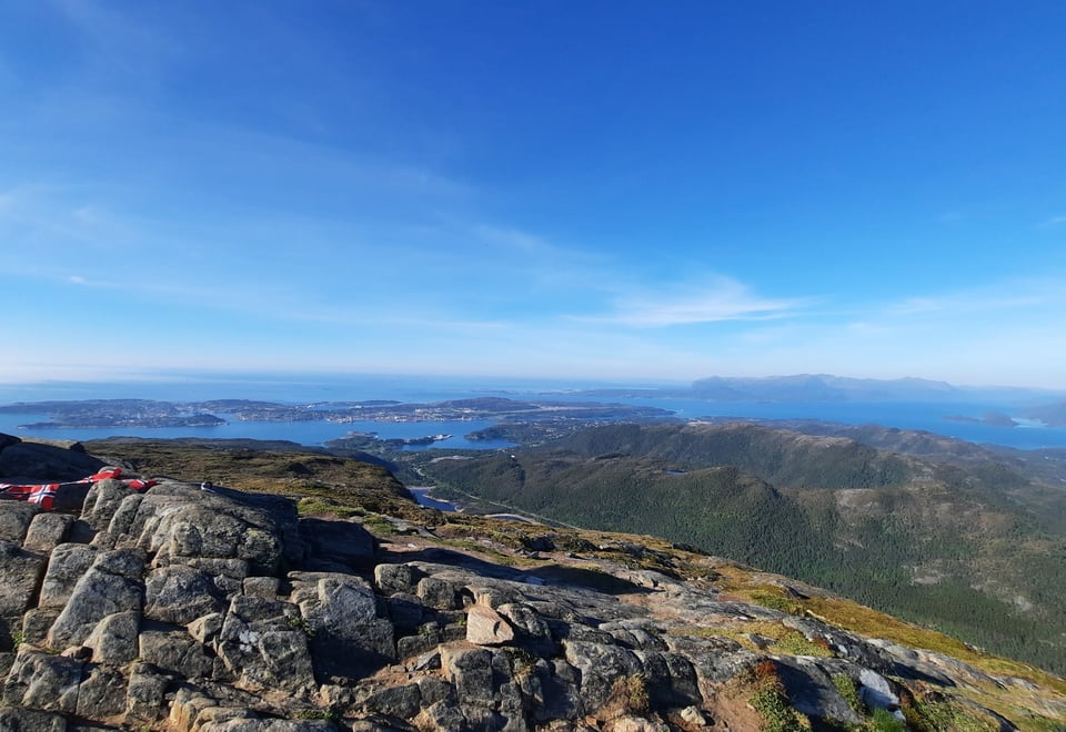 Bildet er tatt fra det som ser ut som en fjelltopp, med utsikt over skog og hav. Det er blå himmel.