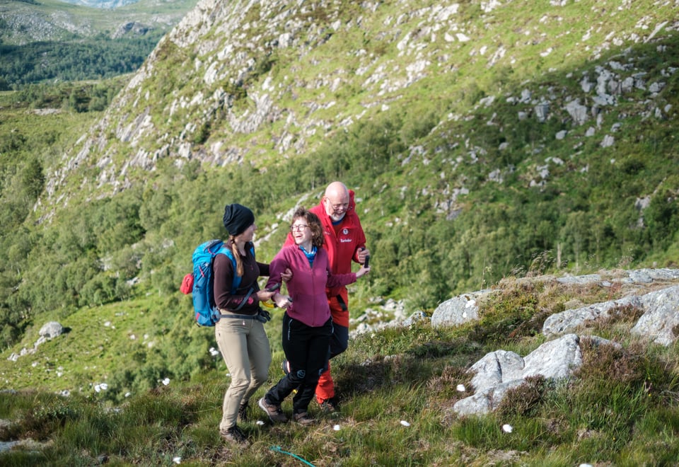 Tre personer går i fjellterreng. Damen i midten har en gul bøyle som hun holder i og blir støttet av to ledsagere. Alle personene smiler og ler.