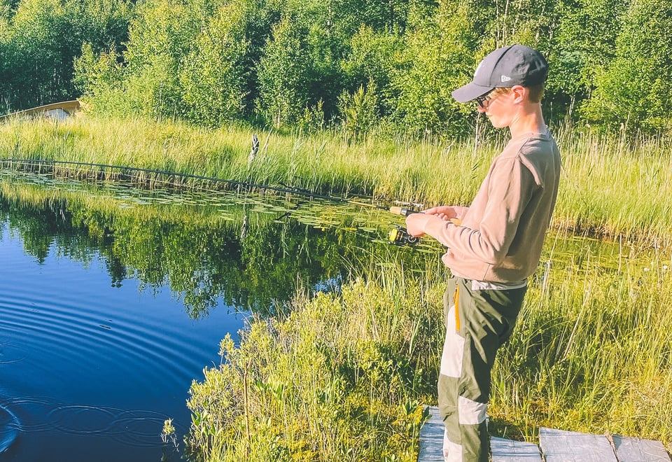 Gutt fisker med stang i et skogstjern. Skog i bakgrunnen, og en skyfrihimmel speiler seg i vannet.