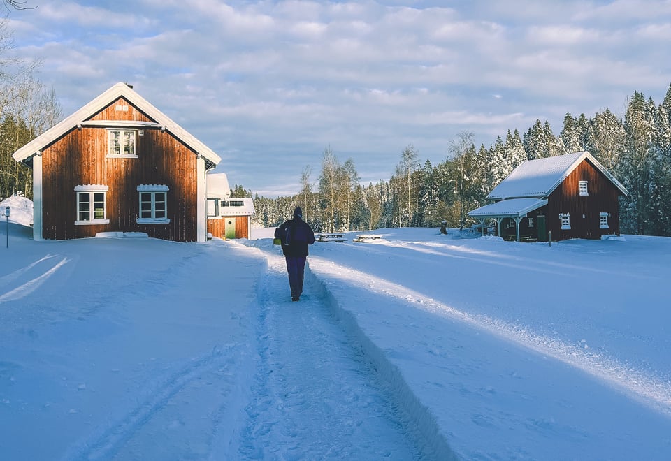 En person går på en snødekt sti mot et rødt trehus, med et annet hus og trær i bakgrunnen under en delvis overskyet himmel.