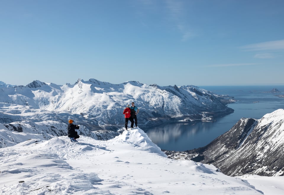 To personer som smiler for kameraet på toppen av en snødekket fjelltopp. Ved siden av er det en person på huk som tar bilde.