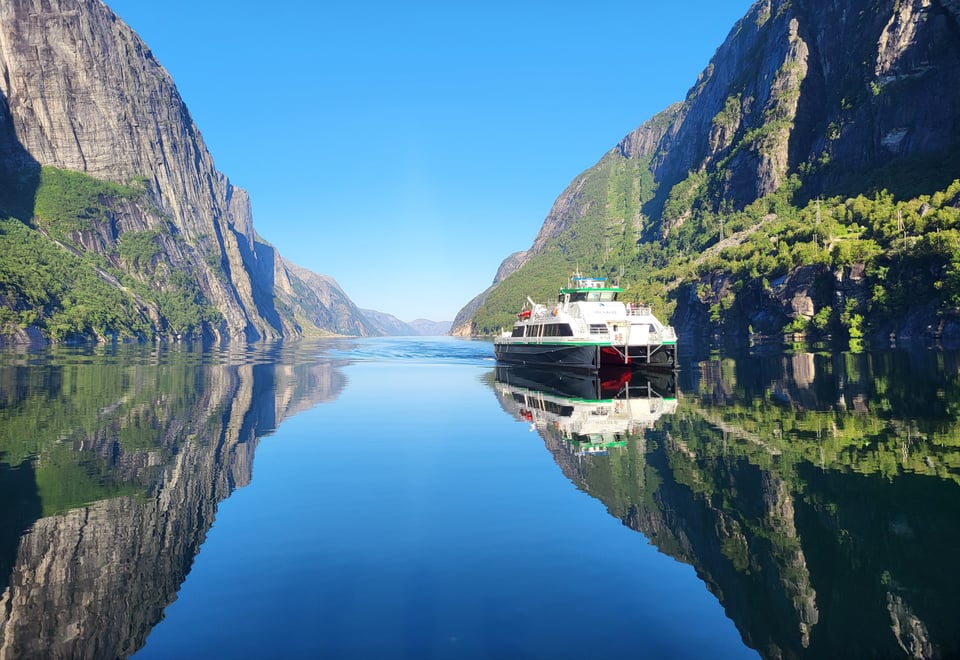 Bilde av en rutebåt som er på vei innover i Lysefjorden. Speilblank fjord med høye fjellsider.
