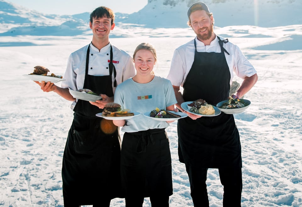 Foto. Utendørs. Tre personer med forkle holder frem tallerkner med mat. Snø og fjell i bakgrunnen.