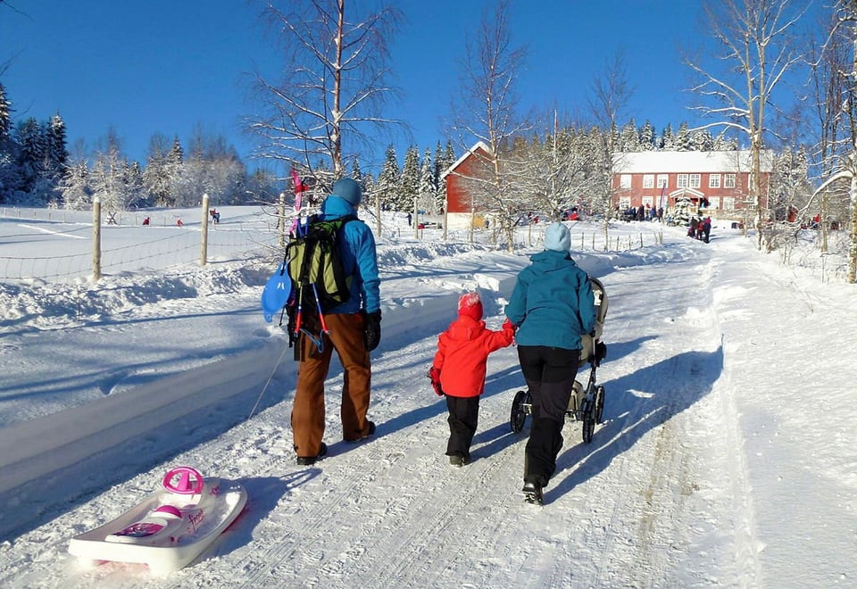 To voksne og et barn triller en barnevogn opp bakken mot en rød gårdsbygning om vinteren, med snødekte bakker rundt. Masse folk er samlet rundt hytta, og noen leker i snøen.