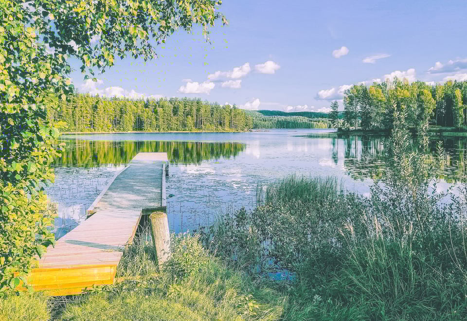 En brygge strekker seg ut i et stille vann, hvor en lettskyet blå himmel speiler seg.
