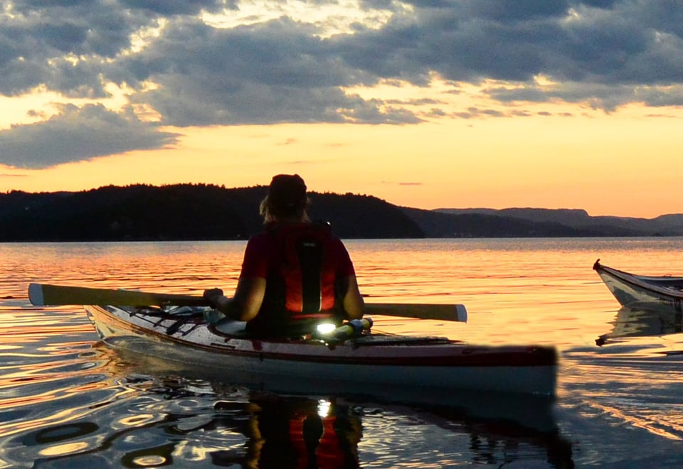 To personer i kajakk padler på stille vann i solnedgangen.