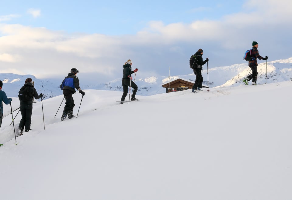 Foto. En rekke med skigåere sett fra siden. Blå himmel, snø og et laftet stabbur i bakgrunnen.