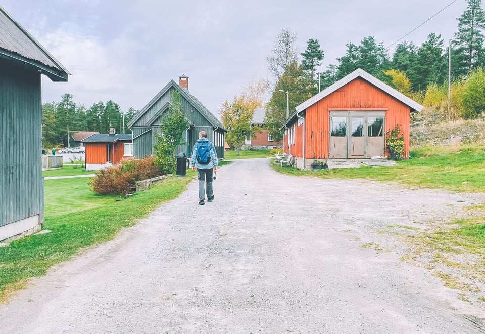 En person med ryggsekk går på en grussti mellom trebygninger, deriblant en rød hytte, i et landlig område med trær og gress.