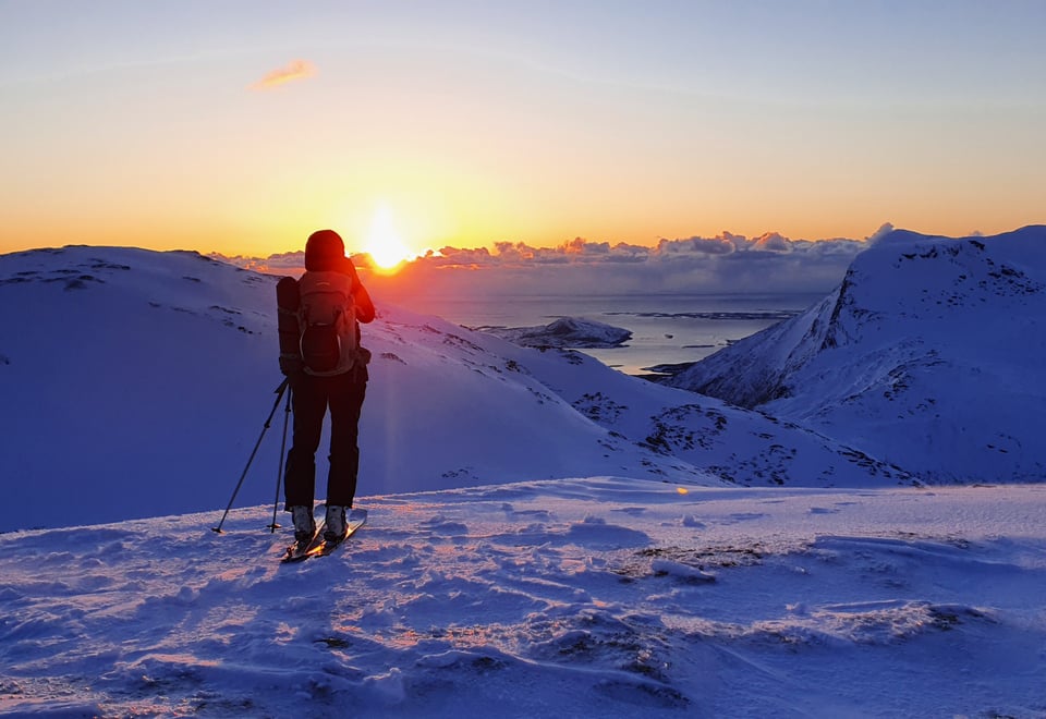Siluetten av en person med topptur utstyr på toppen av en snødekked toppp som ser utover solnedgangen