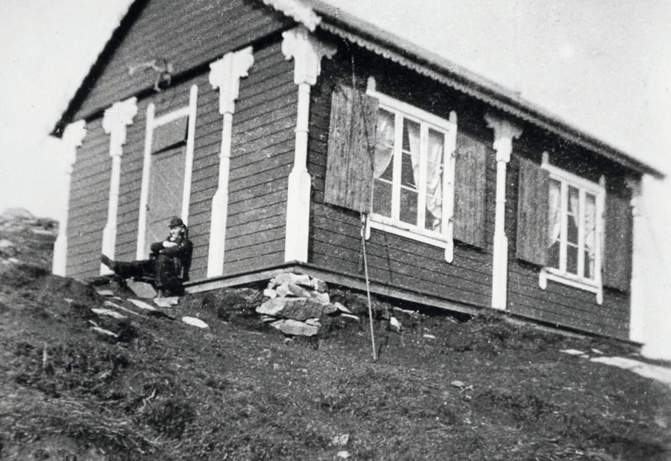 Gammelt svart-hvitt bilde av en liten turisthytte. To vinduer på langsiden, og en inngangsdør på kortsiden. Den står i en skråning. 