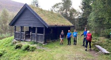 Fire turgåare ved ei brunbeisa enkel hytte med torv på taket 