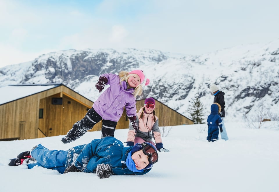 Barn leker i snøen foran en moderne trefarget hytte. Fjell i bakgrunnen.