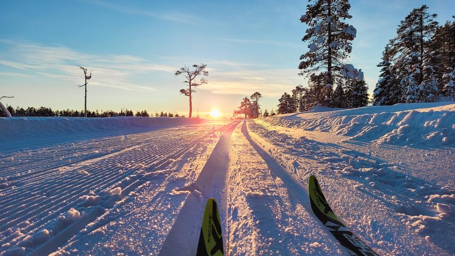 Et oppkjørt og maskinpreparert skispor, sett fra bakken, med et par ski tittende ut fra bunnen av bildet, og en sol lavt på horisonten