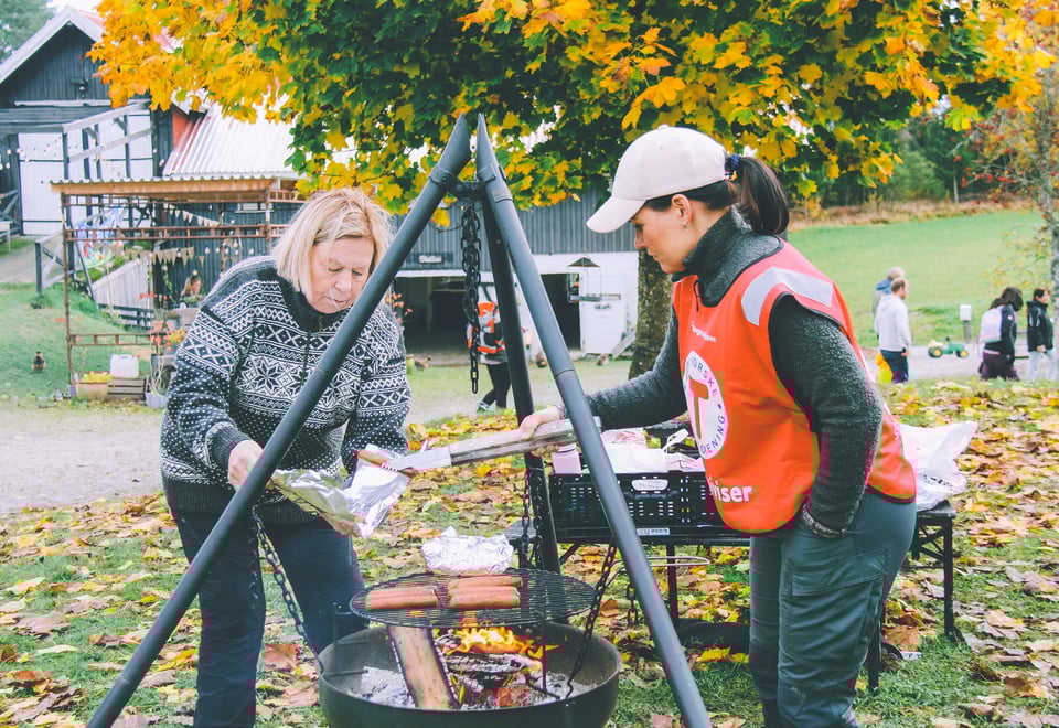 To kvinner står ved en bålpanne under høsttrærne og lager mat over åpen ild, med et gårdshus og andre mennesker synlige i bakgrunnen.