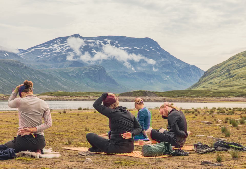 Ungdommer som sitter i yoga-posisjon i et landskap omgitt av fjell og vann.