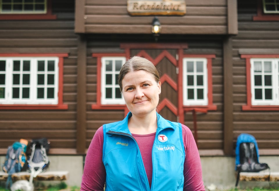 dame med blå vest med t på står på et hyttetun med et bredt smil om munnen 