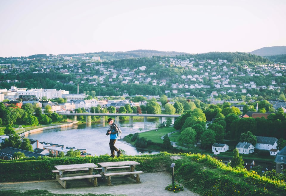 Eldre mann med hviss skjegg og stort speilreflekskamera på rem rundt halsen går forbi oss, med Elgeseter bro og nidelven synlig bak ham, med byen rundt. 