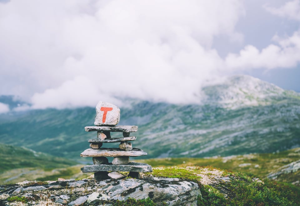 Bildet viser en varde av stablede steiner, med en rød T malt på den øverste steien, stående ute i naturen. Himmelen er overskyet. Fjell i bakgrunnen. 