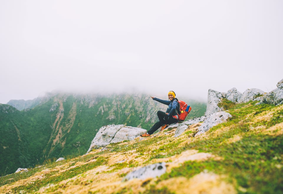 Kvinne sitter på en fjellside og peker, fjelltoppene rundt er dekt av tåke.