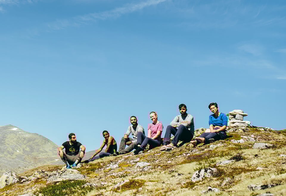 En gruppe mennesker sitter på en fjellrygg, men høye fjell i bakgrunnen.