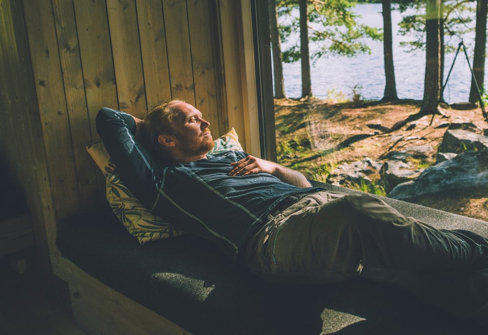 En person hviler på en sofa ved et vindu som vender ut mot en skog og en innsjø. Solstråler skinner gjennom trærne og skaper en fredelig atmosfære. Personen ser avslappet ut og nyter den rolige omgivelsen inne i en hytte