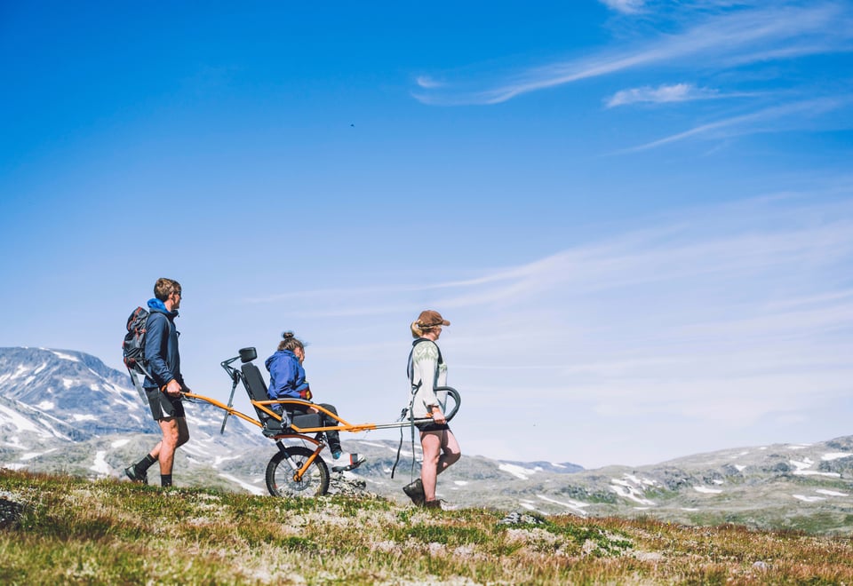 Tre personer ute på tur, på rekke og rad, sett fra siden idet de reiser fra venste mot høyre. Den midterste sitter i en rullestol. Noen fjell i det fjerne. Skyfri himmel.