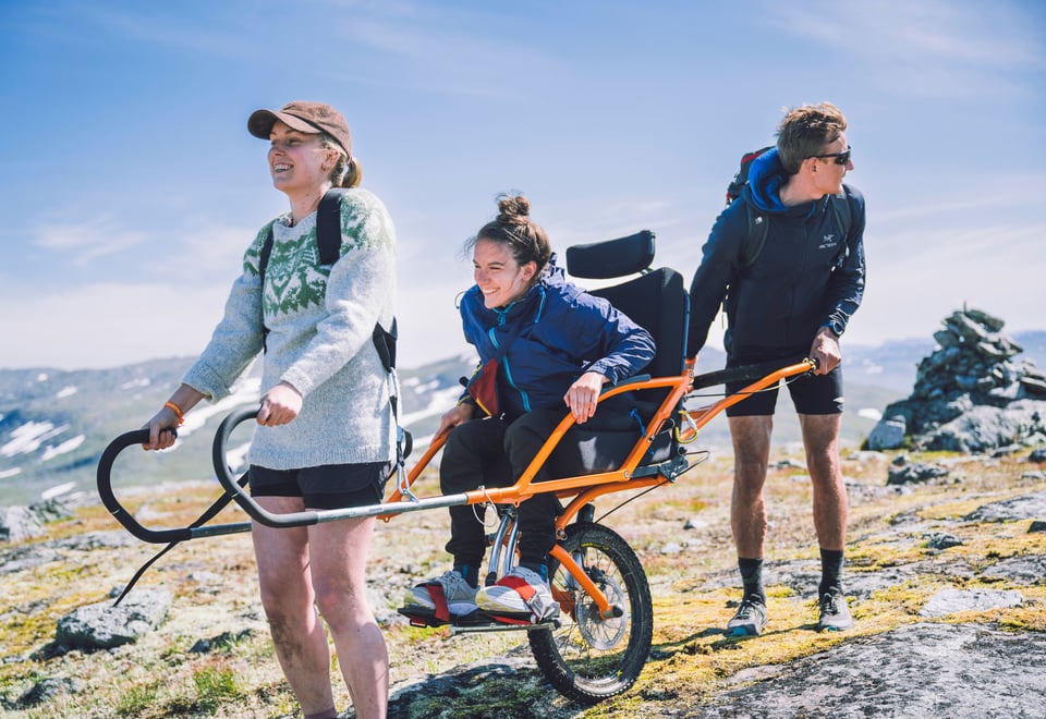 Tre personer ute på tur, der to stykker hjelper den i midten med å holde/bære en modifisert rullestol for bruk ute i naturen. 