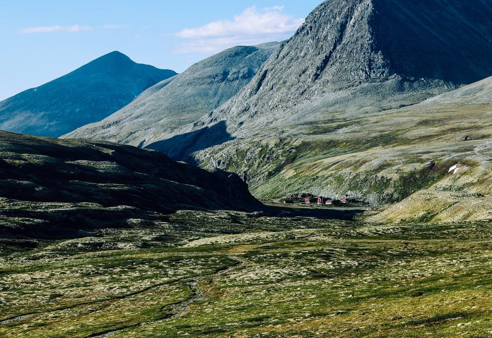 Et fjellandskap med steinete og kuperte fjell som strekker seg mot horisonten. I bunnen av dalen kan man se noen små bygninger som står samlet, omgitt av grønt landskap og lyng. Fjellene i bakgrunnen gir et majestetisk og dramatisk inntrykk, mens noen områder fortsatt har små flekker av snø.