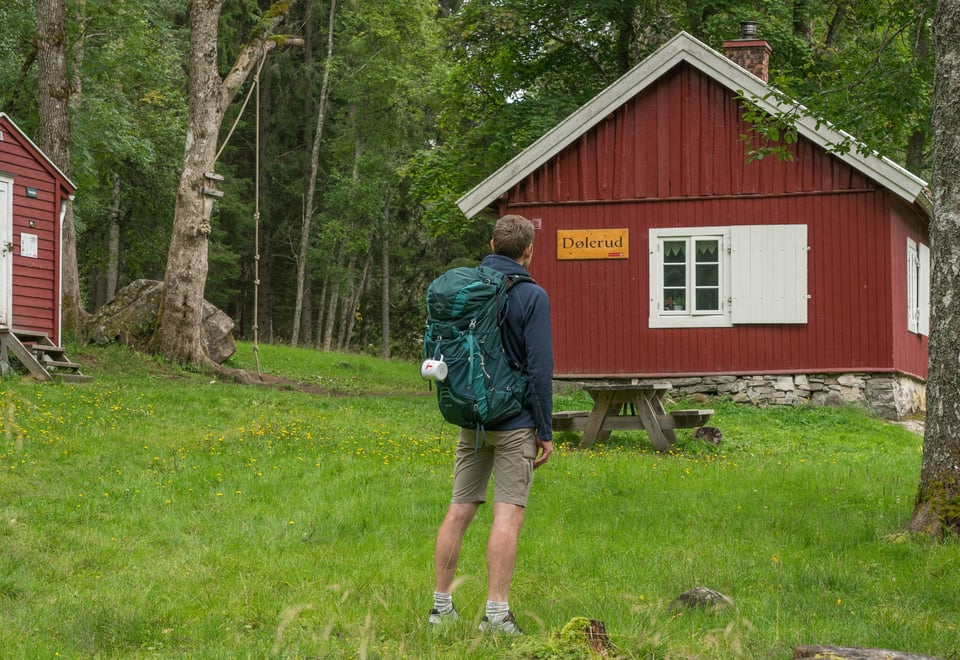 Person med sekk foran en rød hytte. 