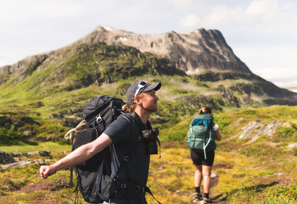 To personer ute på tur, begge med stor tursekk på ryggen. Stort fjell i mellomgrunnen