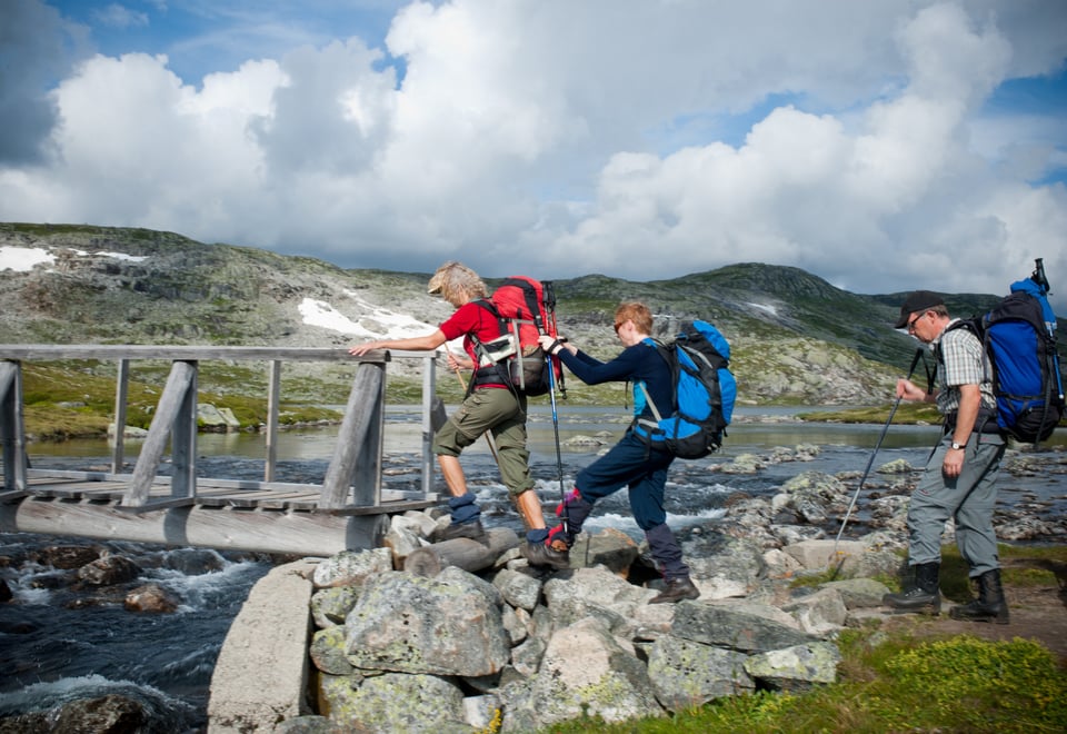 Tre personer med ryggsekk er ute på tur, og er i ferd med å gå ombord på en trebru.