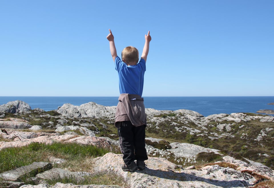 Barn står på fjelltopp med hendene i været og ser mot havet. Foto
