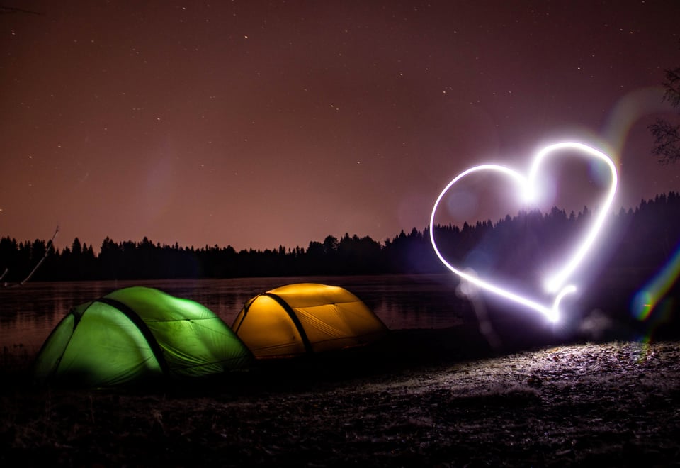 To telt lyser opp i forskjellige farger ved et tjern i skogen. Et stort hjerte laget av lys henger i lufta ved siden av teltene. Det er mørkt, og på himmelen ser man stjerner og lys etter et fly. 