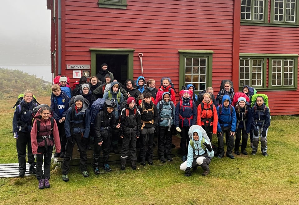 Mange ungdom med jakke og sekk  i står foran en rød hytte med grønne karmer. 