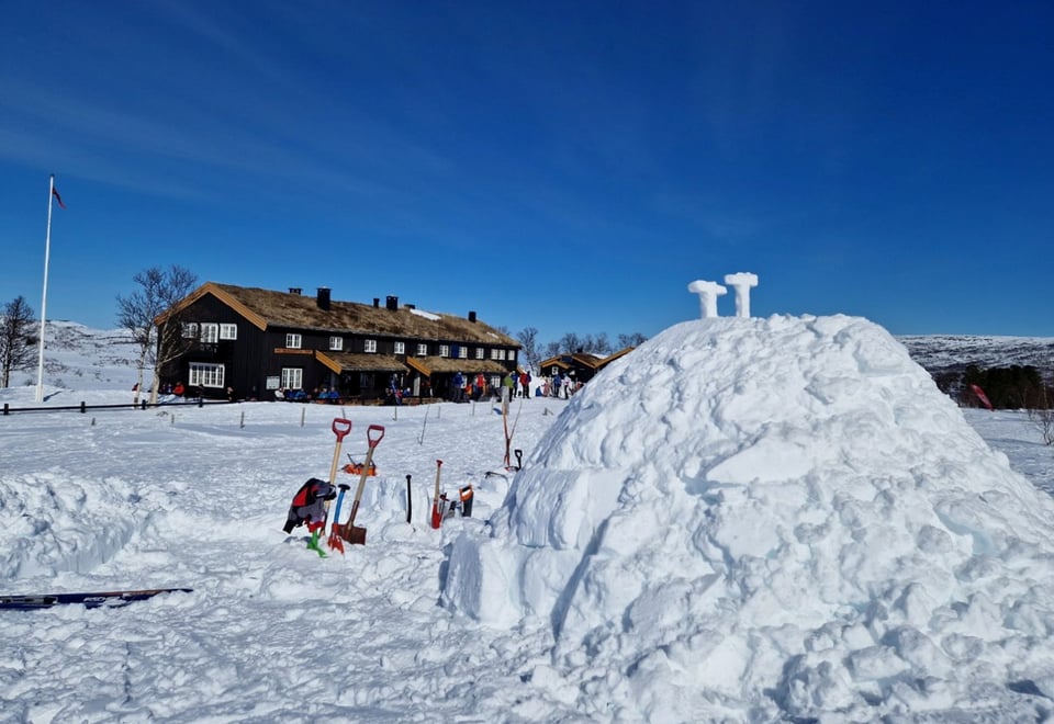 En stor igloo i forgrunnen, med utstyr som spader og sag satt i snøen rundt, og to T-er formet av snø stilt ut på toppen. En lang brun hytte i bakgrunnen, med en gruppe folk vrimlende foran. 