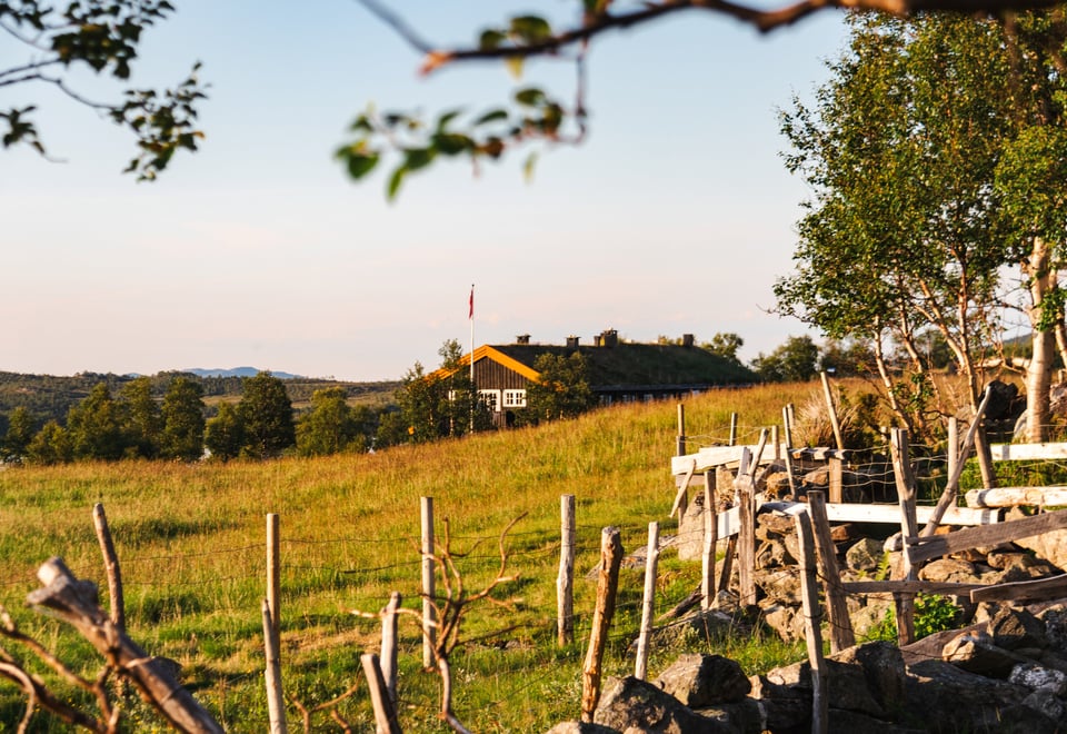 Bildet viser en hytte som ligger på andre siden av en eng. Pent sommervær.