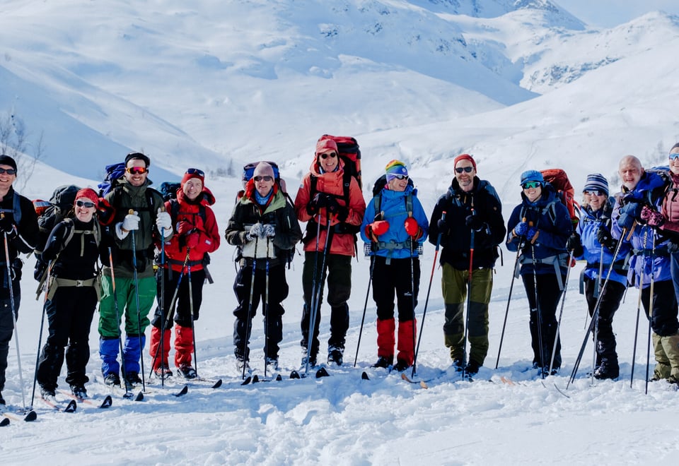 A group of smiling hikers equipped with backpacks and ski poles, dressed in winter gear, stand together on a snow-covered mountain landscape, ready for a winter adventure under clear skies