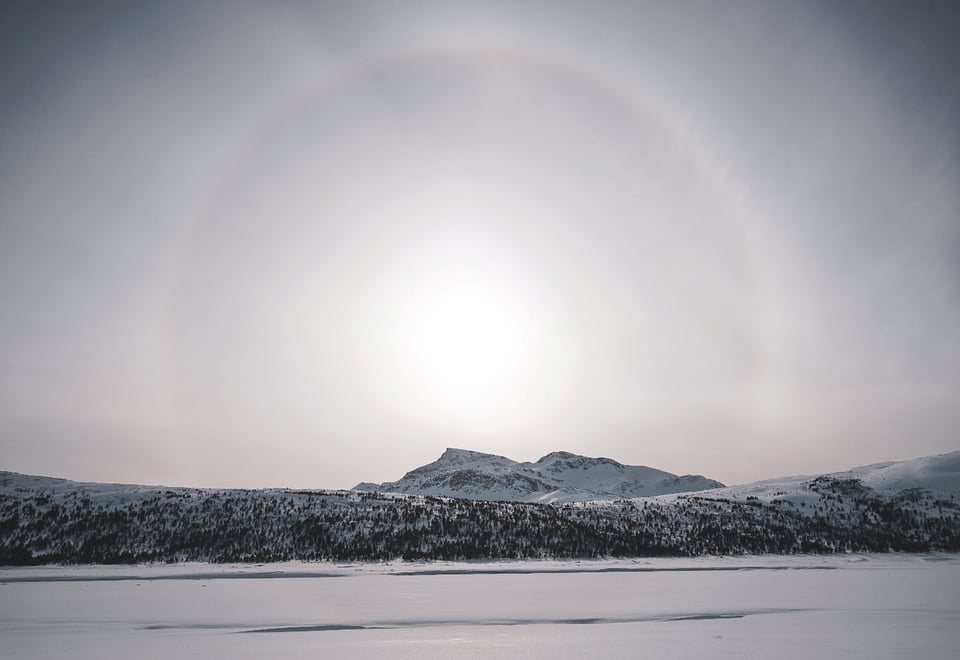  Bildet viser en stor åpen hvit himmel, med en blek sol i midten, omgitt av en ring-halo av lys, med et snødekket fjell rett under, som er del av en fjellkjede som strekker langs horisonten. Snødekket slette i forgrunnen.