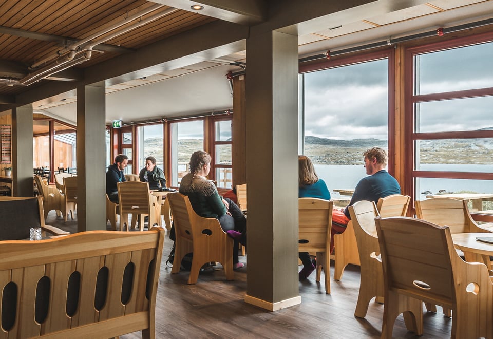 et innendørs kaféområde med store vinduer som gir utsikt over et vakkert fjellandskap. Noen mennesker sitter rundt bordene og prater, mens de ser ut mot naturen utenfor. Kaféen har treinteriør, lyse trestoler og et moderne, men koselig preg. I bakgrunnen kan man se fjell og en innsjø under en overskyet himmel
