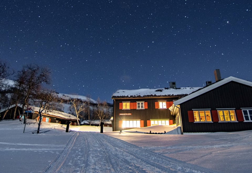 Bildet viser en turisthytte om vinteren, med både landskap og tak dekket av snø. Det er kveld, og lysene er på i de fleste vinduene, med skyfri himmel over.