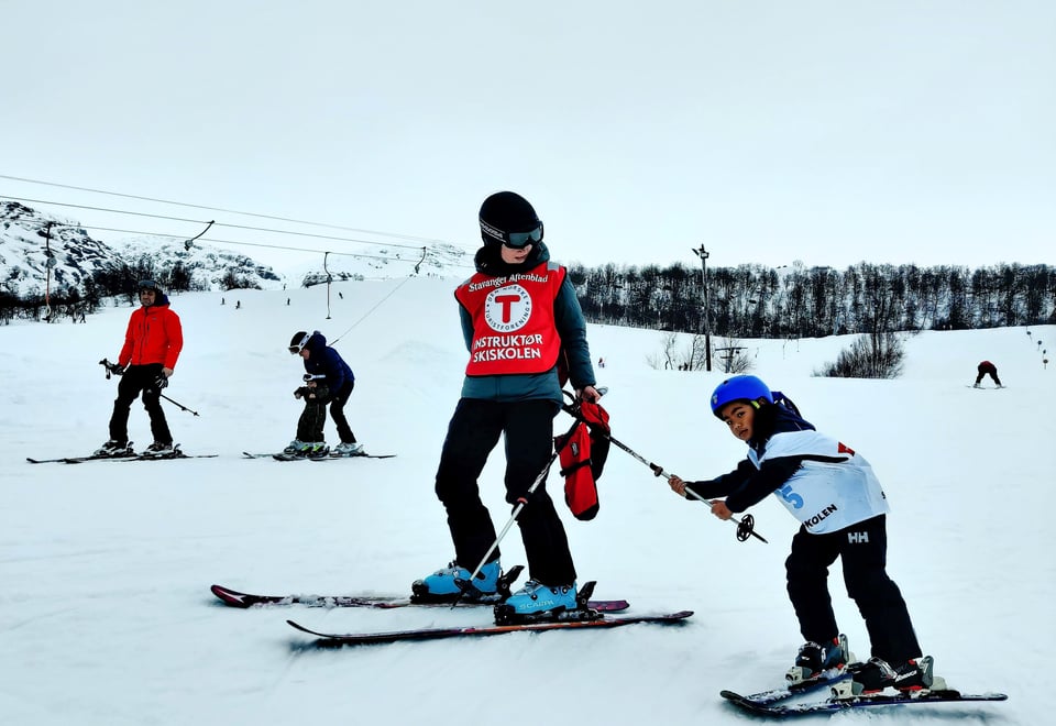 Et barn på slalomski holder seg fast i en skistav som en instruktør holder etter seg.