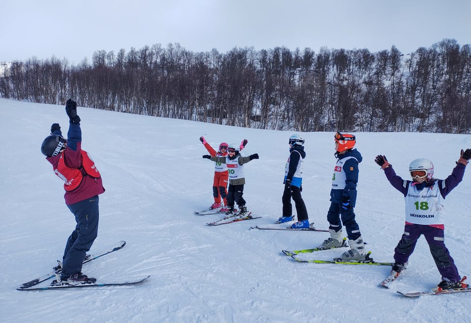 En skiinstruktør står med hendene over hodet og instruerer flere barn med slalomski på beina.